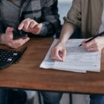 Duas pessoas analisam formulários tributários e fazem cálculos com uma calculadora sobre uma mesa.