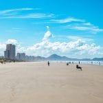 Vista da faixa de areia da Praia Grande, com o mar, montanhas e a paisagem urbana ao fundo.