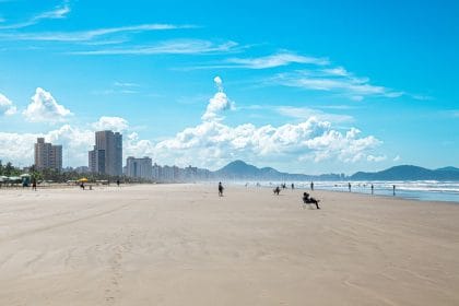Vista da faixa de areia da Praia Grande, com o mar, montanhas e a paisagem urbana ao fundo.