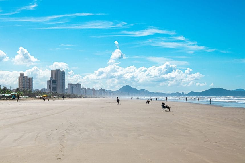 Vista da faixa de areia da Praia Grande, com o mar, montanhas e a paisagem urbana ao fundo.