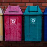 Quatro contêineres coloridos para separação de metal, plástico, vidro e papel diante de uma parede de tijolos.