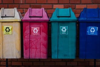 Quatro contêineres coloridos para separação de metal, plástico, vidro e papel diante de uma parede de tijolos.