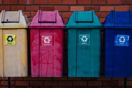 Quatro contêineres coloridos para separação de metal, plástico, vidro e papel diante de uma parede de tijolos.