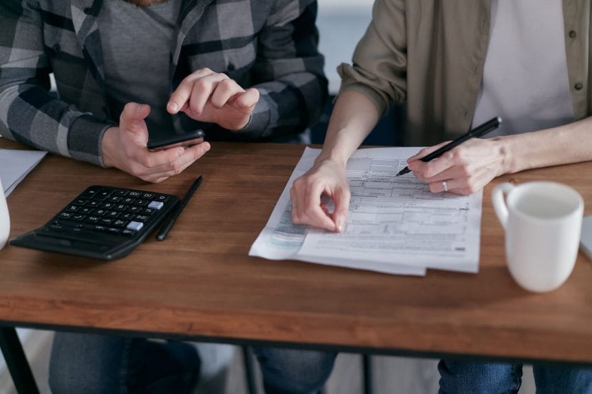 Duas pessoas analisam formulários tributários e fazem cálculos com uma calculadora sobre uma mesa.