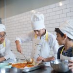 Alunos aprendem a preparar pizza, espalhando molho sobre a massa em uma cozinha.