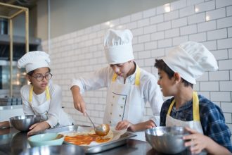 Alunos aprendem a preparar pizza, espalhando molho sobre a massa em uma cozinha.