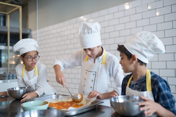 Alunos aprendem a preparar pizza, espalhando molho sobre a massa em uma cozinha.