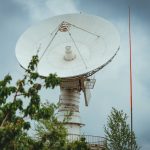 Antena parabólica de comunicação instalada em uma área cercada por vegetação, sob céu nublado.