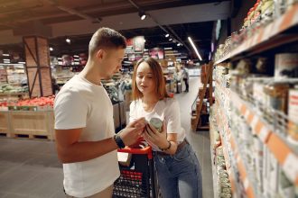 Casal compara um produto diante das prateleiras de um supermercado enquanto empurra um carrinho de compras.